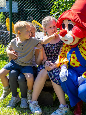 Familotel Maskottchen Happy sitzt mit Kinder zusammen auf einer Bank im Freien